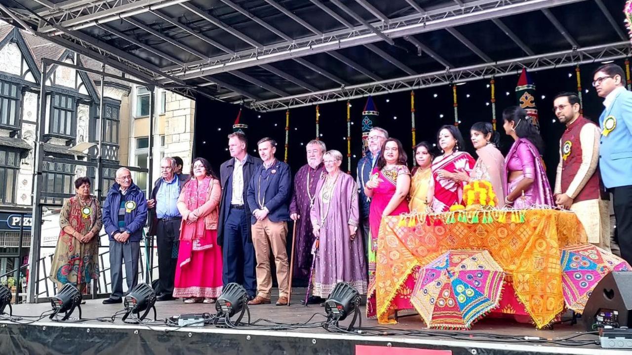 A large group of people, standing on a stage, with a table, with brightly covered cloth on it.