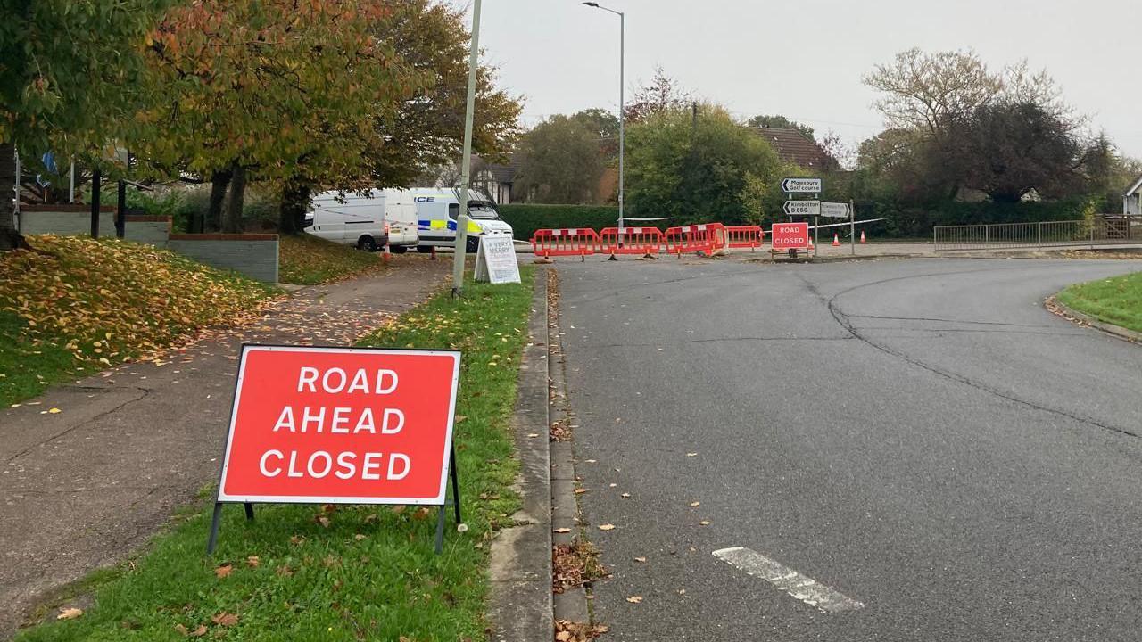 A road closure in Bedford, with a red road closed sign, a cordon in the distance, a road and a path. A police van is in the distance, three road signs and another white van. Leaves are on the path.