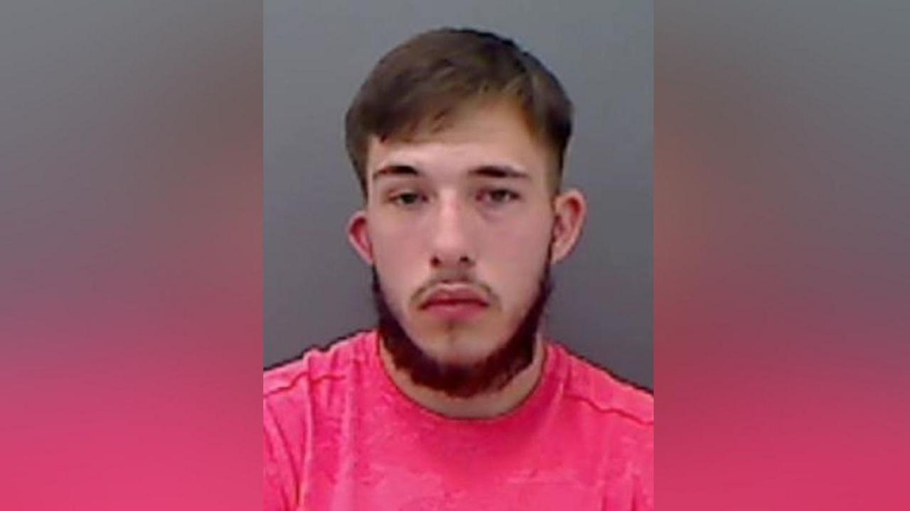 Mugshot of a man in a bright pink t-shirt with a dark beard and hair.