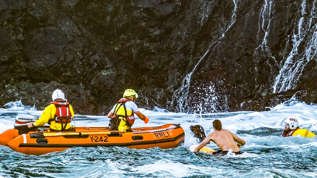 The picture shows a sea rescue operation involving the RNLI. Three rescuers are in action - one in the water wearing yellow and red RNLI life jackets, and two in an orange inflatable rescue boat marked Y242 along with a paddleboarder.