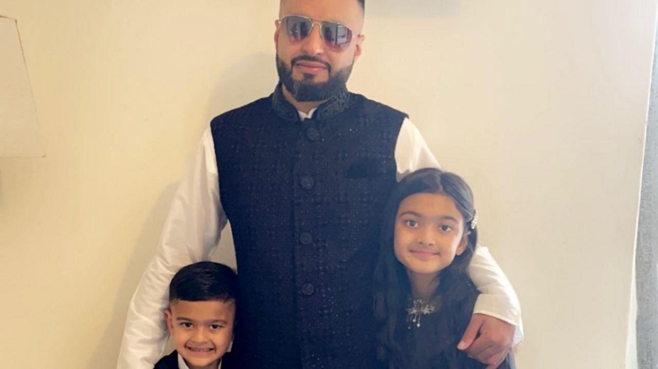 A man in dark glasses stands smiling with his arms around a young boy and girl.