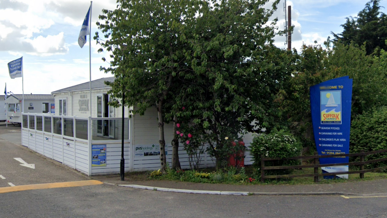 The entrance to a holiday park with a blue sign reading "Welcome to Suffolk Sands", and flags running down the driveway. 