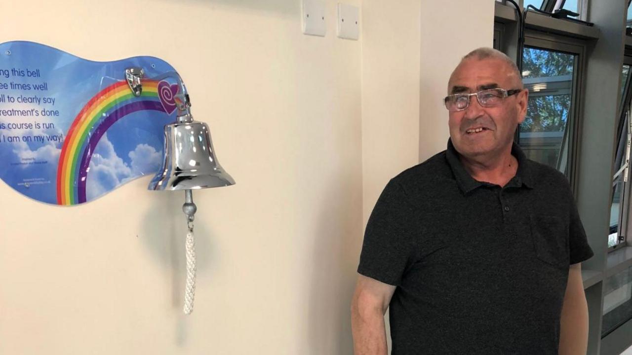Peter Firth in a black top with short hair and glasses looking towards a silver bell on a beige wall. The bell is mounted on a poster with a rainbow and sky scene with a positive message. The bell has a rope on it.