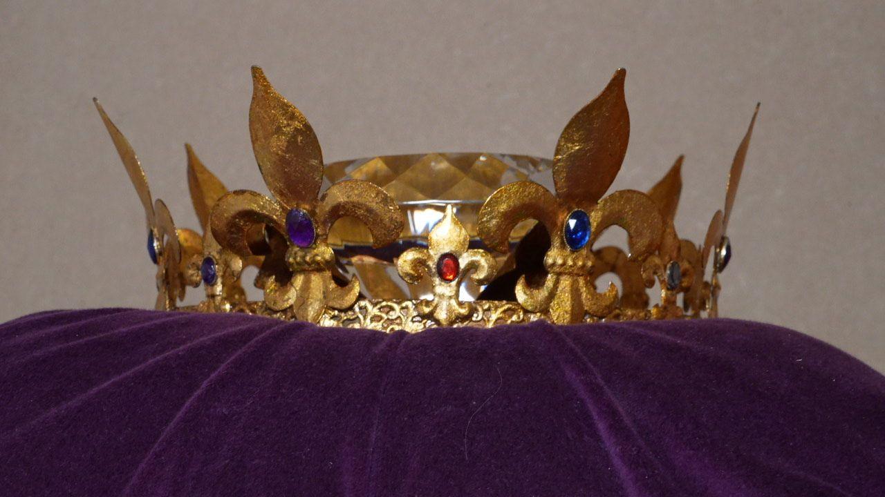 Golden crown with fleur-de-lys motifs around the crown's base. The upright fleur-de-lys are each decorated with a blue stone. In the middle of the crown's head lies an enormous diamond, made of paste.