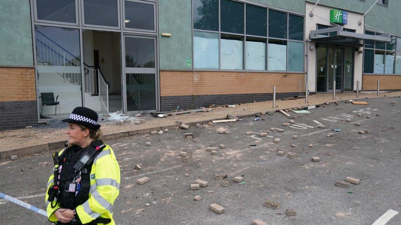A woman in police uniform is on the bottom left of the photo. A sign is above a door on the right and rubble is in the road.