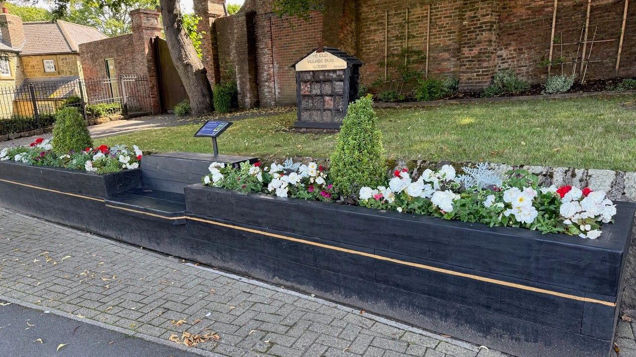 A long wooden bench which is painted black and gold. There is seating in the middles and either said are flower beds filled with colourful plants. There is a stone pavement to the front and grass at the back.