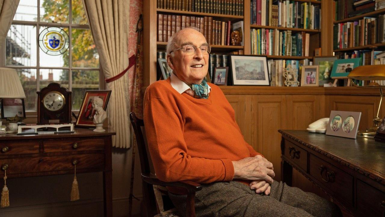 Geoffrey Bond sitting in an office in a period house wearing a bright orange jumper 