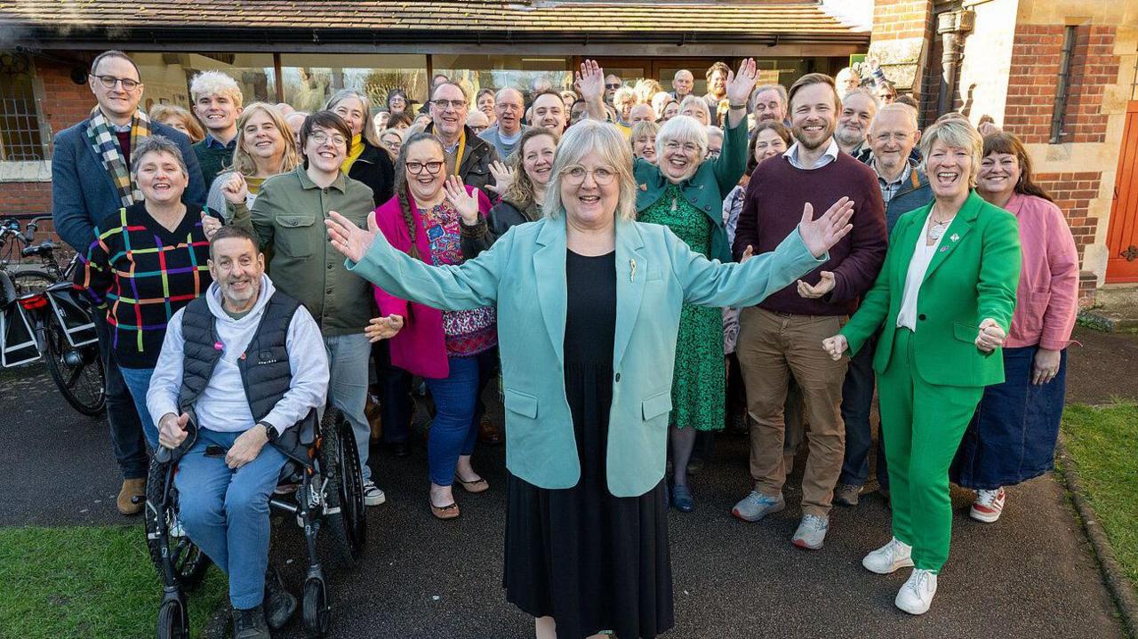 Members of the Liberal Democrats standing in a group smiling for camera- with Lorna in a mint colour jacket and black dress standing in front of the group with her hands spread out 