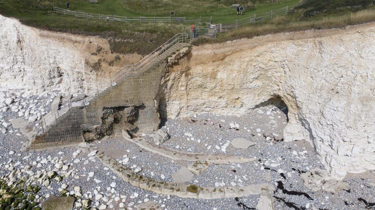 An aerial image of brown stone steps leading down a white chalk cliff face. A person in a red top is stood at the top of the steps. Below the cliffs is a pebble beach with large boulders strewn across it. On top of the cliff is green grass.