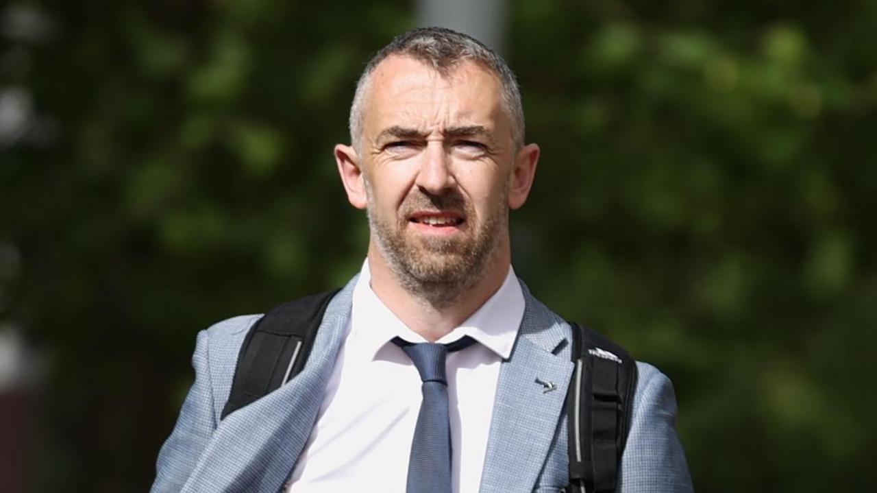 Daithí McKay is wearing a white shirt and a navy tie. He has a grey blazer on and a black rucksack. He has short grey hair and a beard.