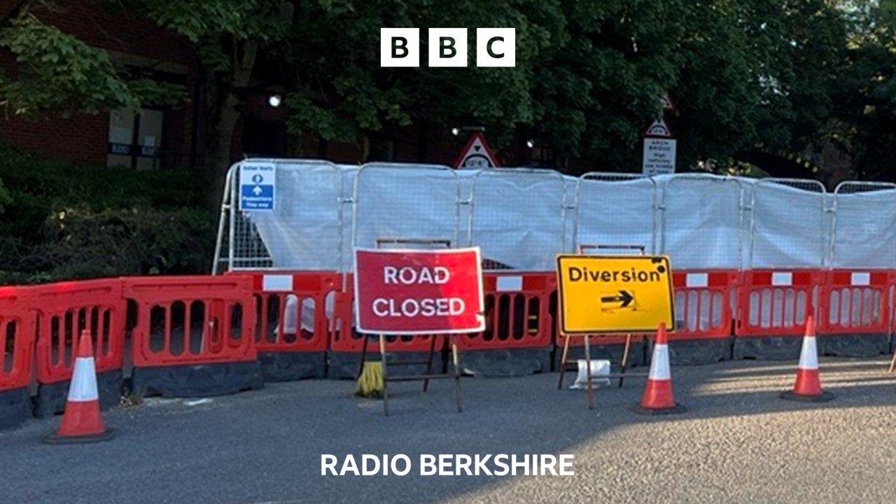 A road closed sign and yellow diversion sign stand in front of red plastic fencing.