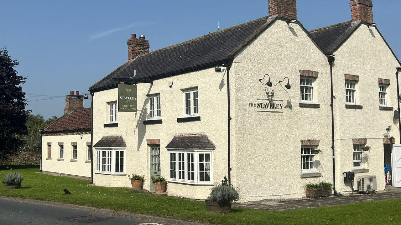 The exterior of a cream-coloured country pub with white windows and a green door. A sign hangs on the front reading 'The Staveley Arms'.