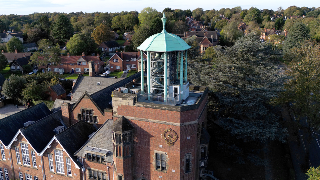 An aerial view of a brown brick building with a central clock tower which has a blue metal structure on top with a bell inside