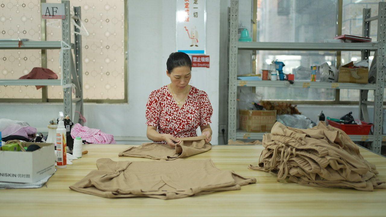 A woman in a red dress stands at a large wooden table folding brown t-shirts. A oile of folded brown trousers also sits on the table. 