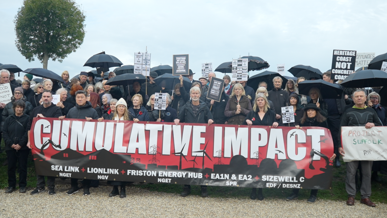 Dozens of people all wearing black and some carrying umbrellas stand in a large group in front of the camera. Some people hold a large banner that reads: "Cumulative impact". The banner has a list of energy projects happening and planned in Suffolk.