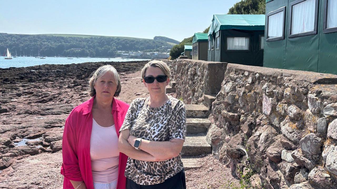 Hut owner Becky Lingard, on the left, wearing a pink cardigan and pink top. Cllr Kate Ewert, on the right, wearing a leopard print top and watch. They are stood on the beach.  There are green huts in the top right corner of the image. In the background in the top left the sea is visible. The sky is blue. 