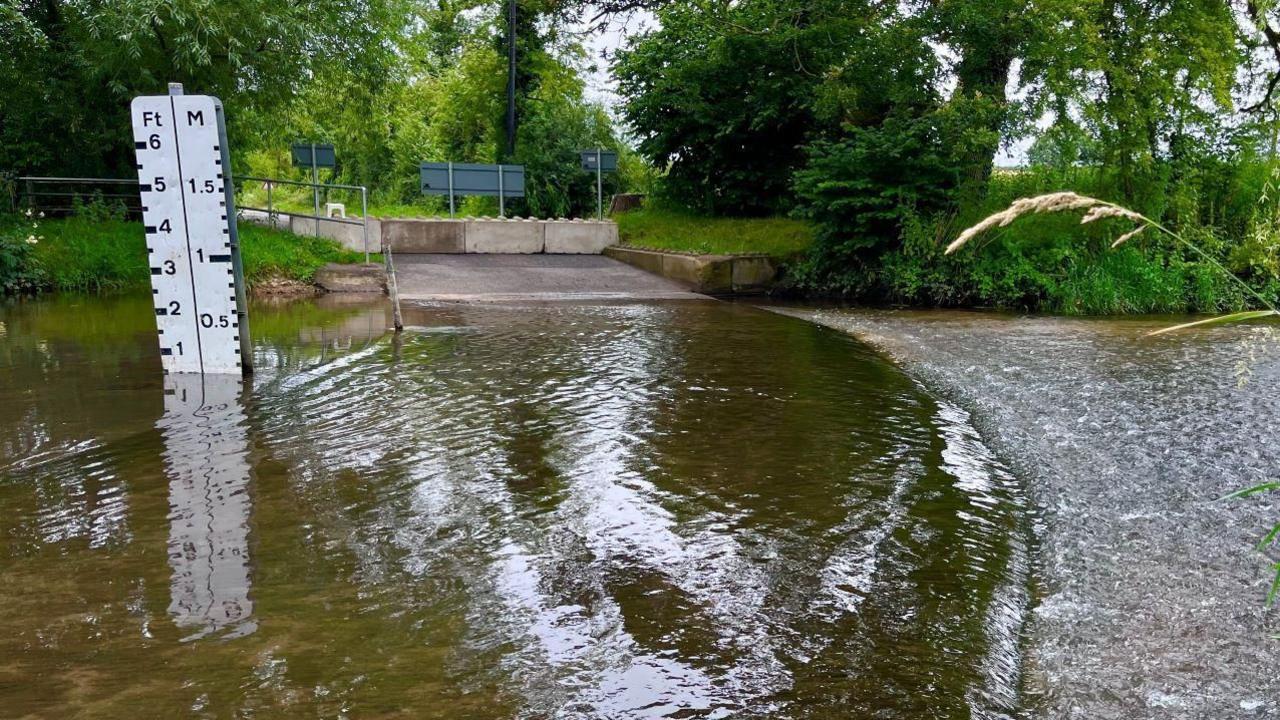 A view of Shotesham ford. It shows a large body of water moving over the road. Barricades can be seen at the end of the road while a water gauge sits on the left hand side showing the depth of the water.