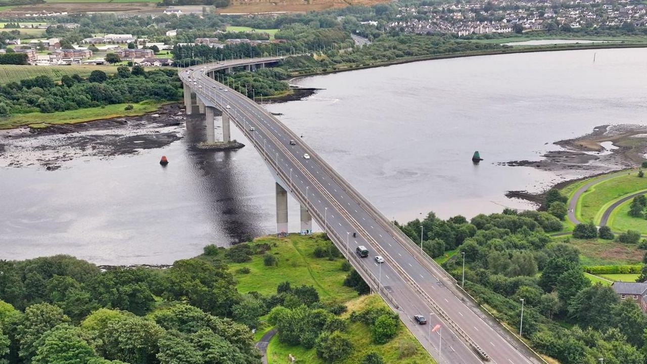 An aerial view of Foyle Bridge. A large grey bridge between two grassy land masses. A number of cars are visible crossing the bridge over the River Foyle.