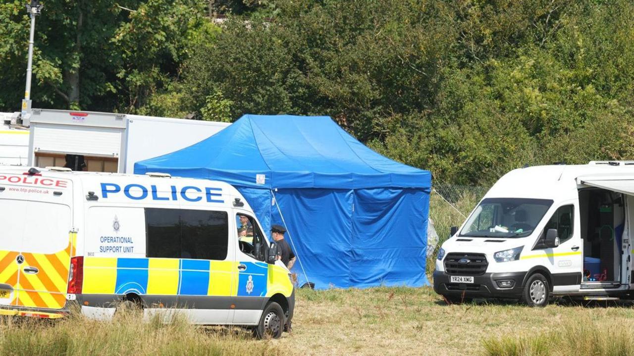 A blue tent stands beside police and support vehicles near the Lewes derailment site. Emergency and investigation teams are working in a grassy area next to trees and railway infrastructure.