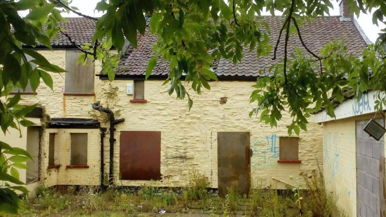 The derelict pub photographed from inside the courtyard. It shows a yellowing building with boarded up windows and doors, and an overgrown patio.