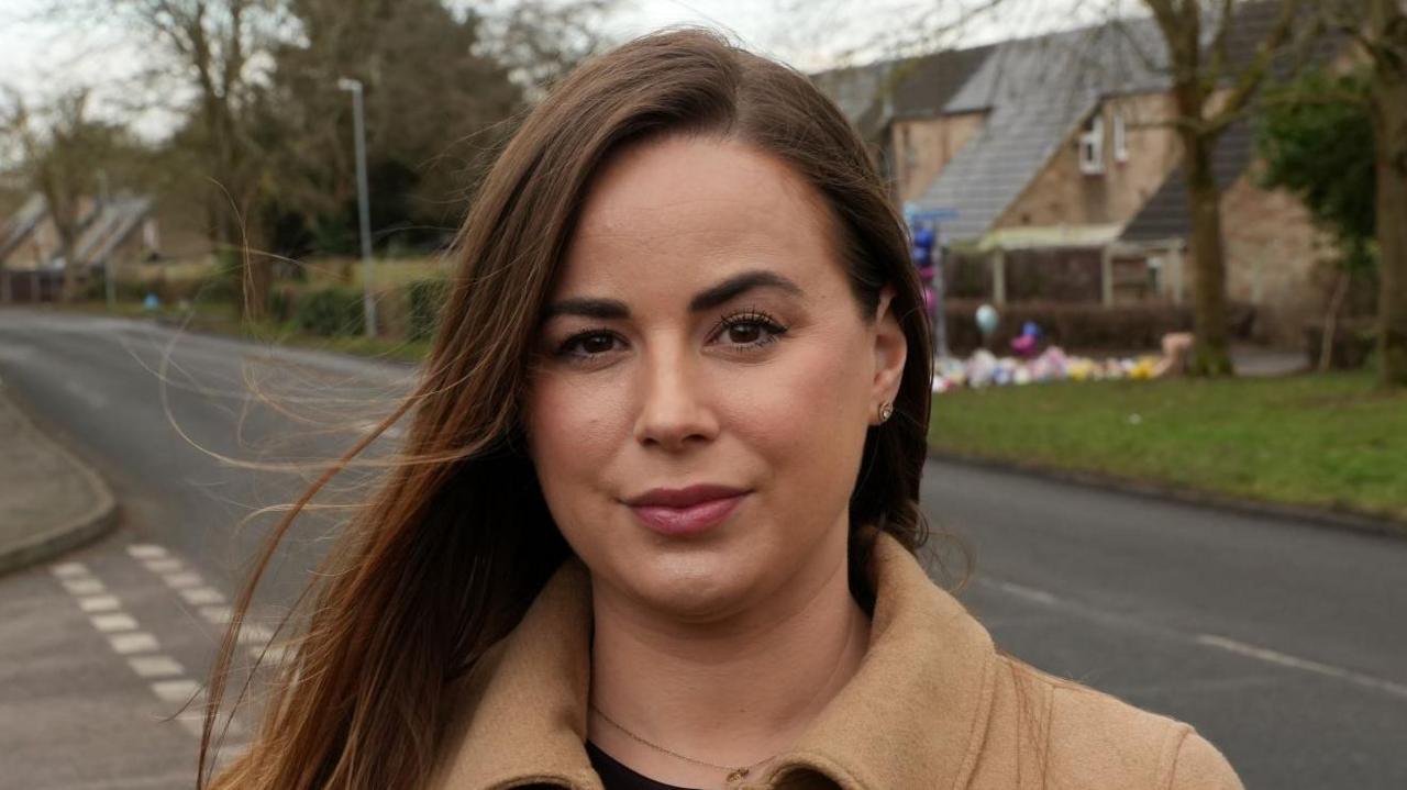 Emma Callaghan standing at the scene of the crash, which is a pedestrian road with flowers and balloons in the background. Emma has long brown hair and is wearing a tan coat with a serious expression on her face.