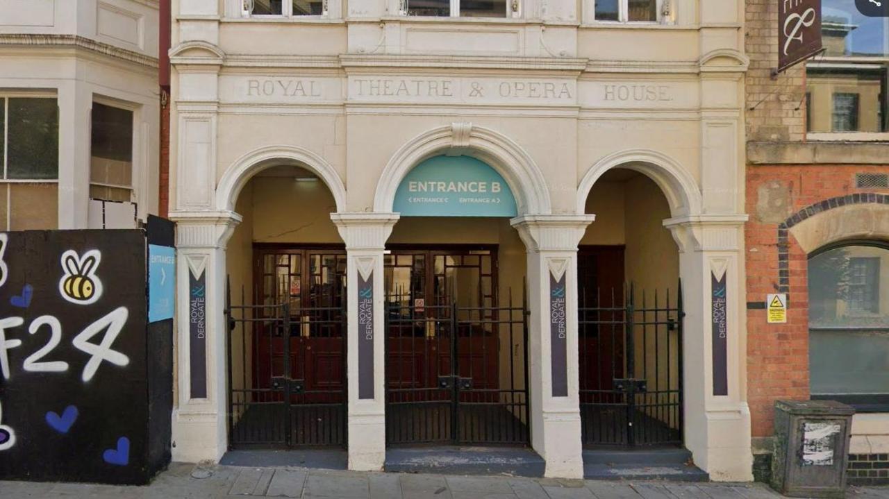Neo-classical entrance to Royal Theatre, showing three cream-coloured arched doorways.The words ROYAL THEATRE AND OPERA HOUSE are engraved above the doors and one of the archways says it is ENTRANCE B. There is a hoarding to the left with F24 visible in white writing.