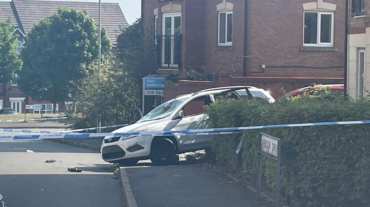 Blue and white police tape is in the foreground, near to an Arbour Drive road sign by a hedge on the right on the photo. A white car is in the centre of the image, with buildings in the background.