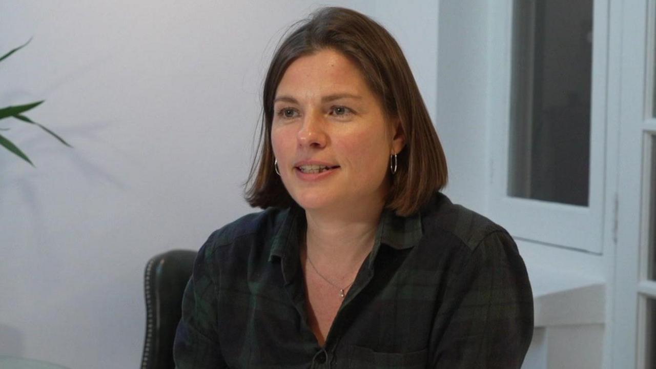 Victoria Wells is seen with short brown hair wearing a dark checked shirt sits at a dining table in a bright room, speaking during an interview.
