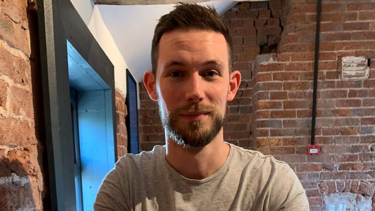 A man in a grey t-shirt with a brown beard standing in a room with brick walls