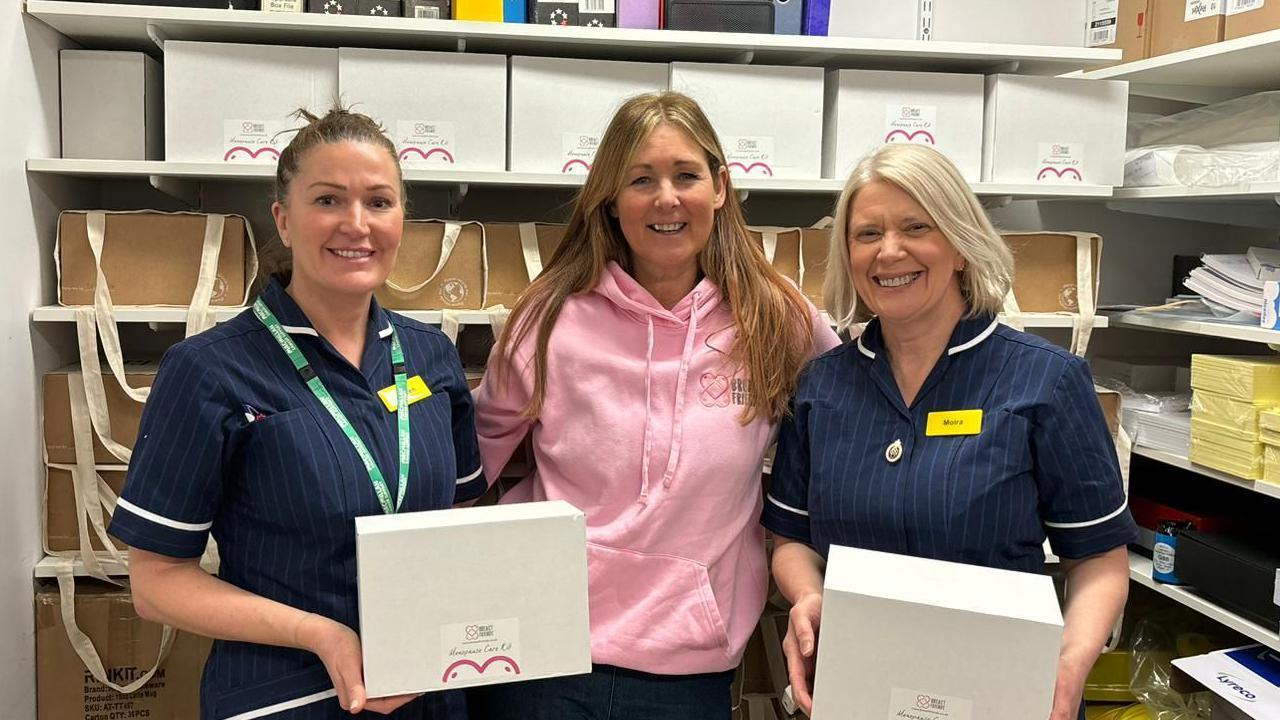 A woman with long brown hair wearing a pink hoodie standing between of two nurses holding white boxes