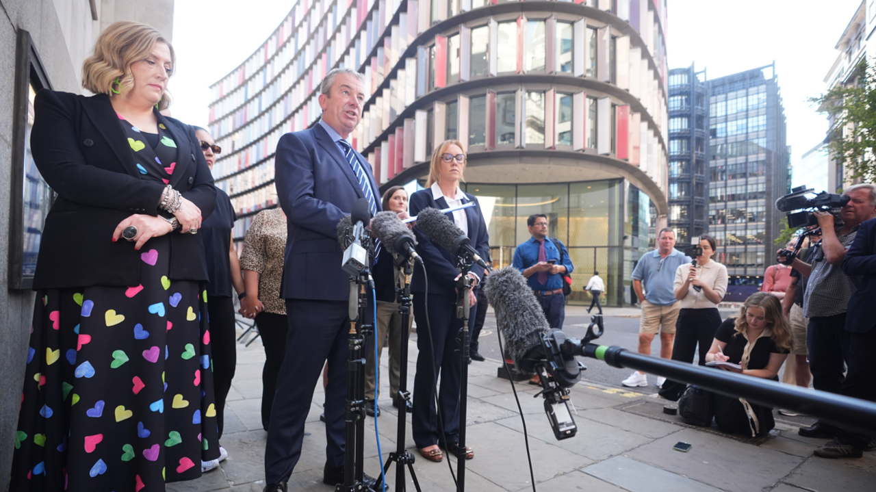 People stand behind microphones on a city street as a speaker addresses reporters outside a modern office building, while journalists and camera crews gather nearby.