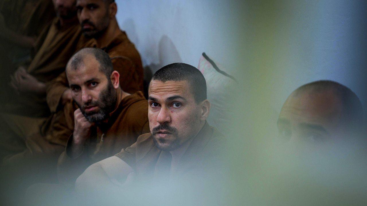 Six male inmates sit together in a row against a wall, looking in the same direction, all dressed in the same brown clothing