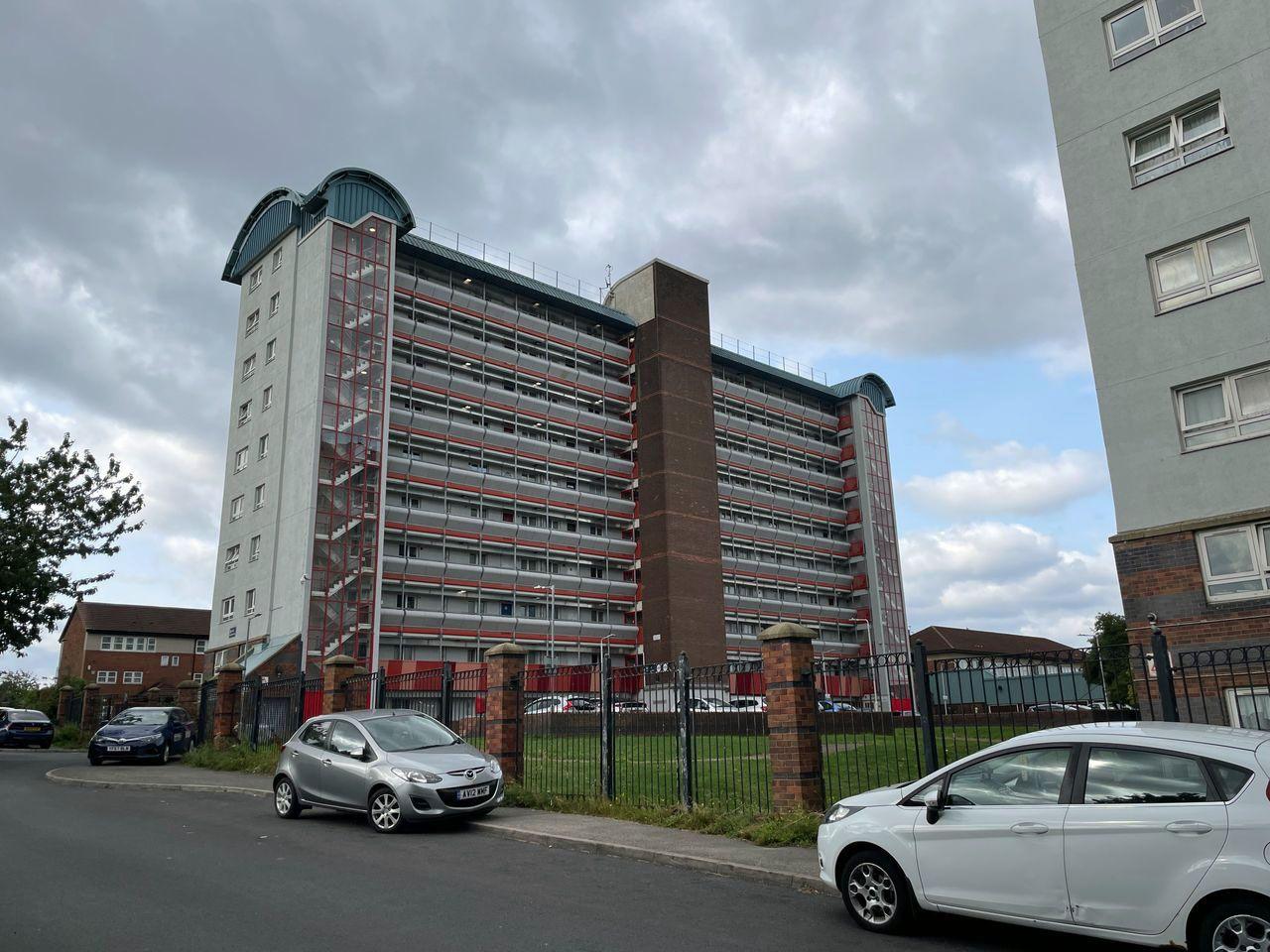 The block of flats in Leeds where Exodus died. Long balconies stretch between three main pillars, you can see cars parked outside and grass.