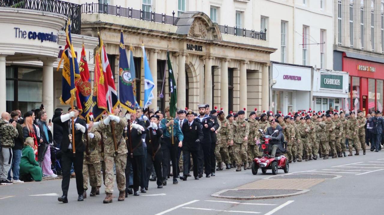 A column of people in military uniform marching along a street, with flags being held by the first few rows. There are tall buildings behind them, some with shop fronts