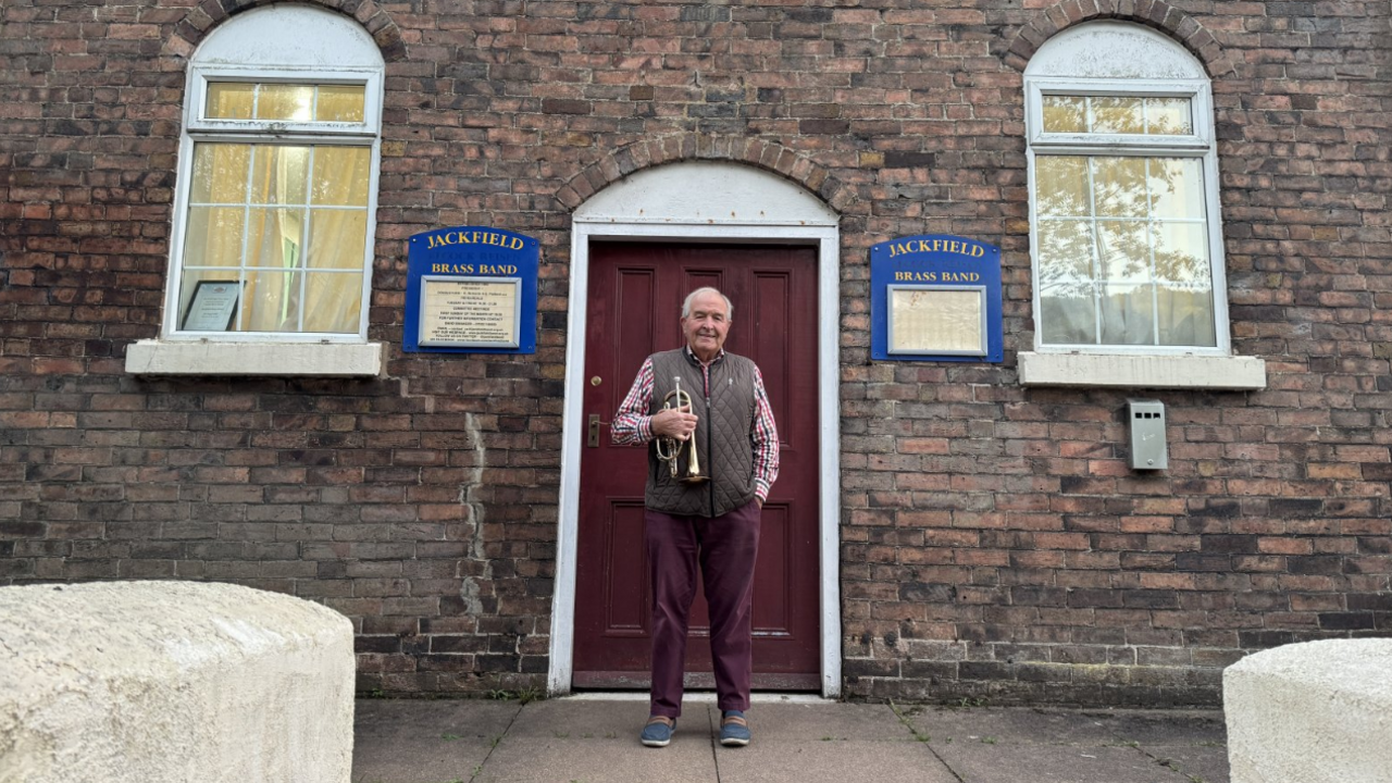 Mr France stands outside the chapel with his cornet. The building is made of bricks with two large windows either side of the maroon wooden door. Mr France is wearing purple trousers, a pink shirt and a brown body warmer. Two blue signs with the name of the band are visible either side of the front door.