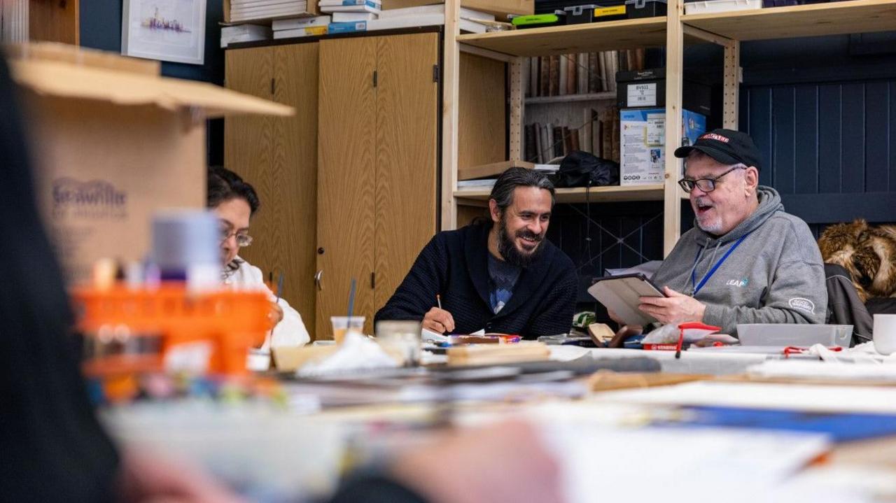 People taking part in a workshop. Two men and a woman are sitting round a table with craft materials on.