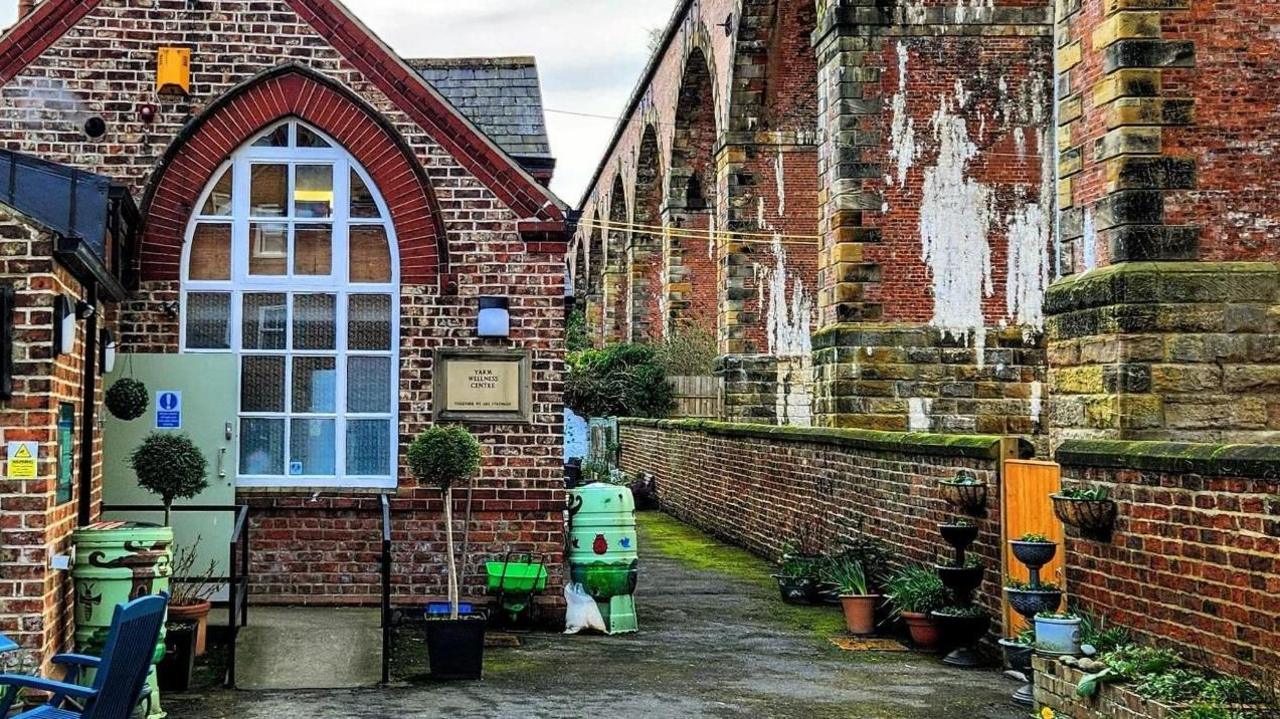 A brick building with a large white arch window. There are planters on a concrete floor. There are large stone arches to the right behind a brick wall.