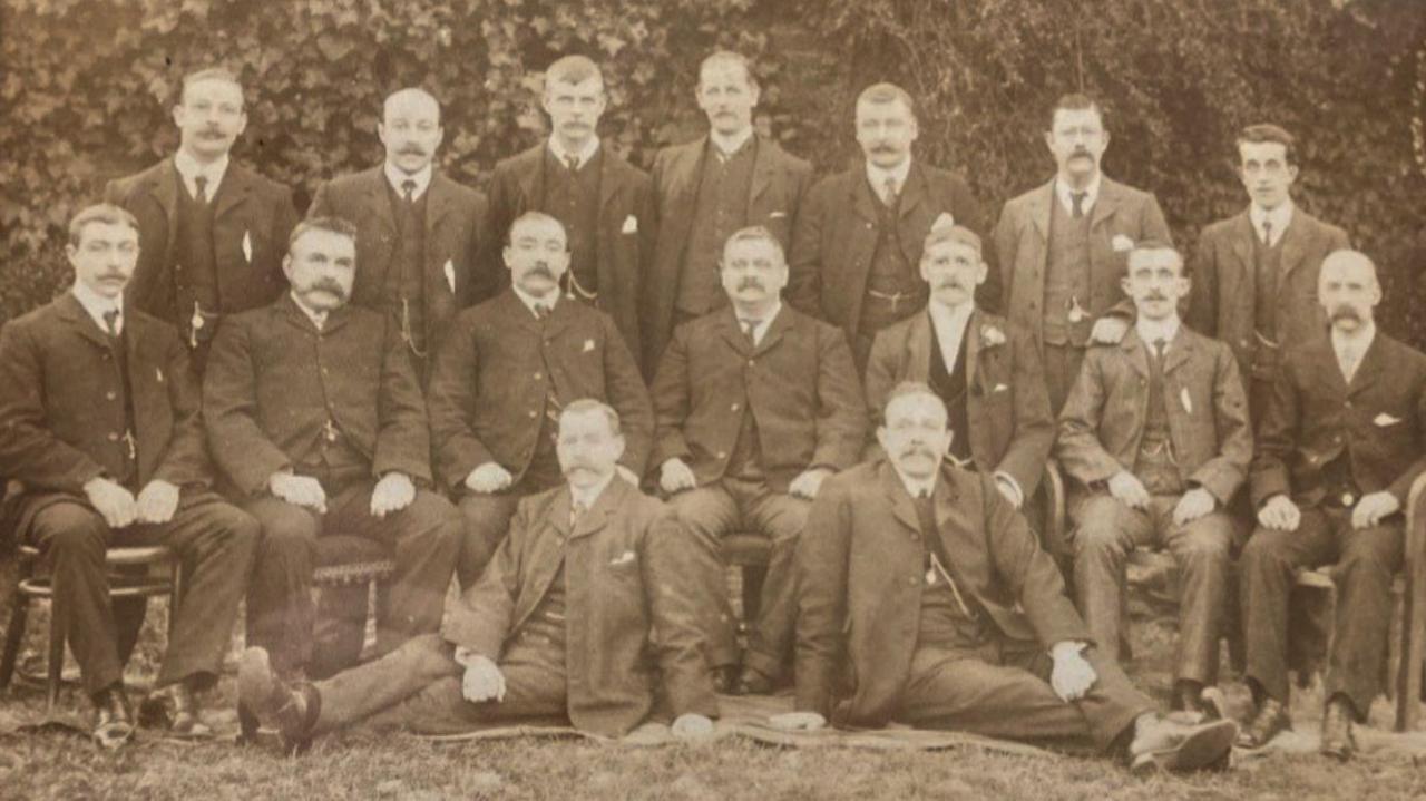 A black-and-white archive photo showing 16 men wearing dark formal suits. Seven are standing in the back row, seven are sitting in the middle row, and two are reclining on the ground at the front.