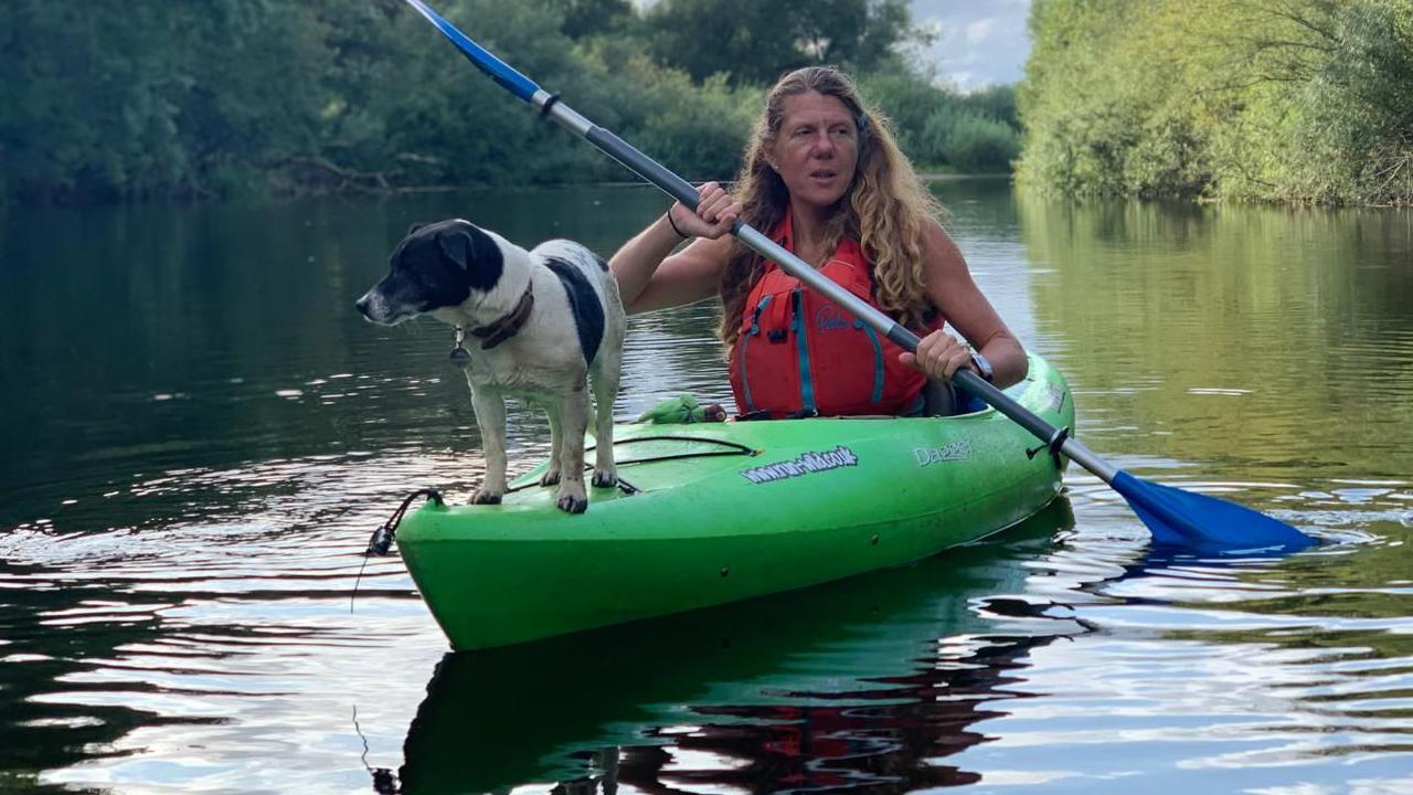 Angela Jones in a green kayak on a river, with a dog stood to the end of it. Trees can be seen on the banks of the river in the background.