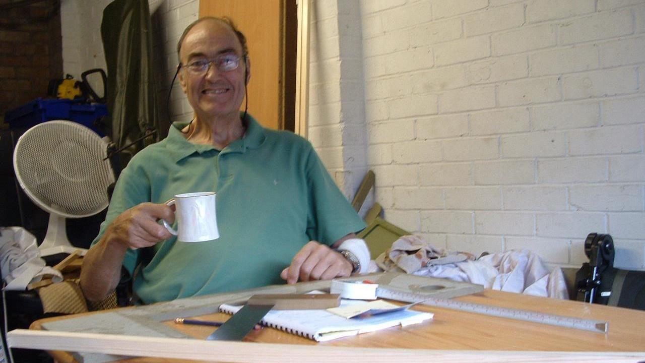 A older man wearing a green polo shirt sits at his work bench, which is covered in tools and equipment. He is holding a white mug and smiling.