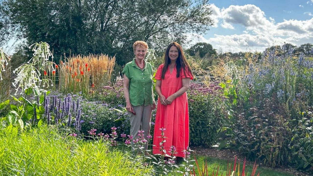 Two women smiling into the camera stood in a beautiful, full and green garden. There are purple, pink, blue and orange flowers around them and a tree behind. It is a sunny day with some clouds in the sky.