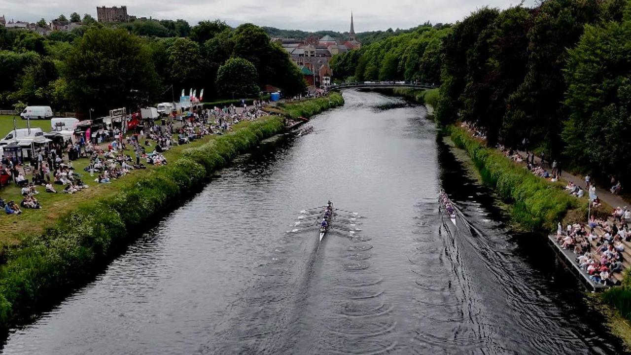 An aerial view of the Durham Regatta. Two rowing boats are on the River with corwds of people watching from the banks. The skyline of Durham City is in the background.