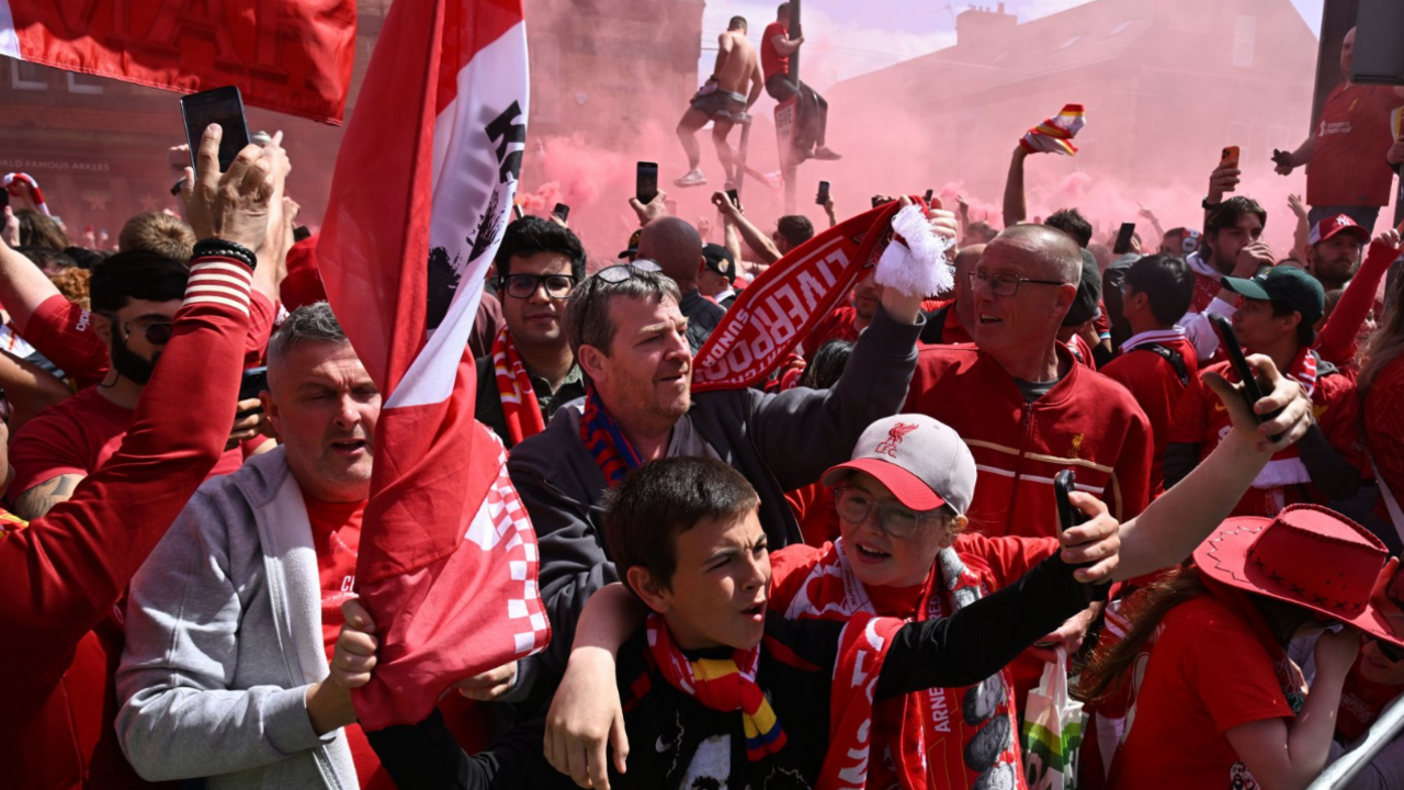 Crowds of Liverpool fans. Many are in the red of the Liverpool shirt, there are flares around them going off so the picture is filled with red smoke.