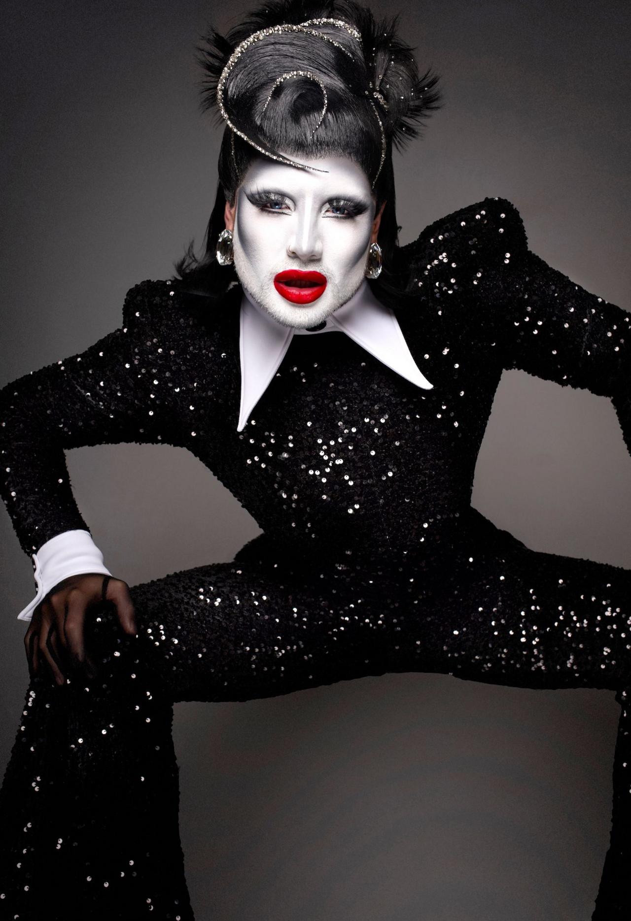 A drag queen with a white face and a sparkly black jump suit poses for the camera, wearing bright red lip stick and a black feathered wig.