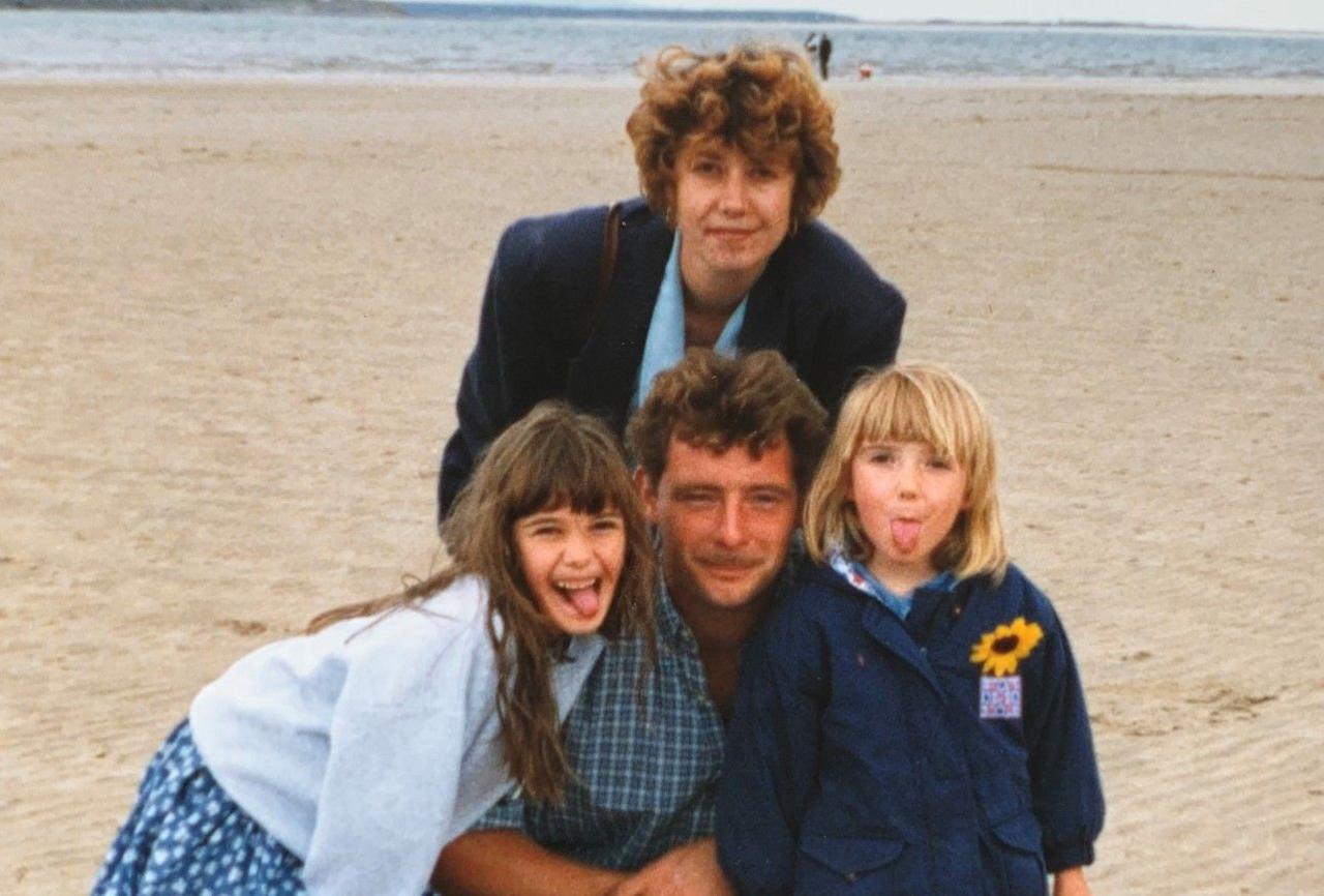 A man is next to two children, with a woman behind them. They are on a beach, with water visible behind that.