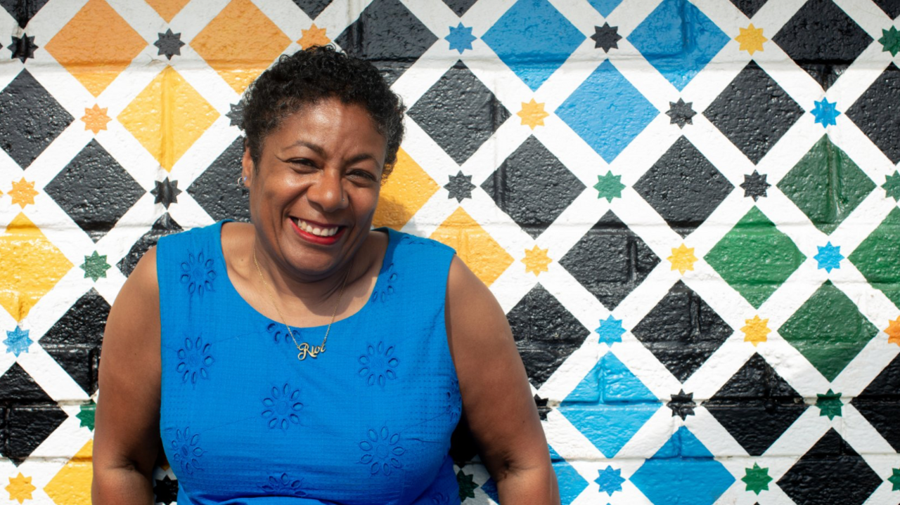 Patrice Lawrence wearing a blue dress sits in front of a colourful background.