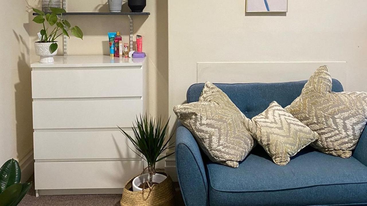 Inside the renovated centre is a small blue couch with golden patterned pillows, plants and a set of drawers. 