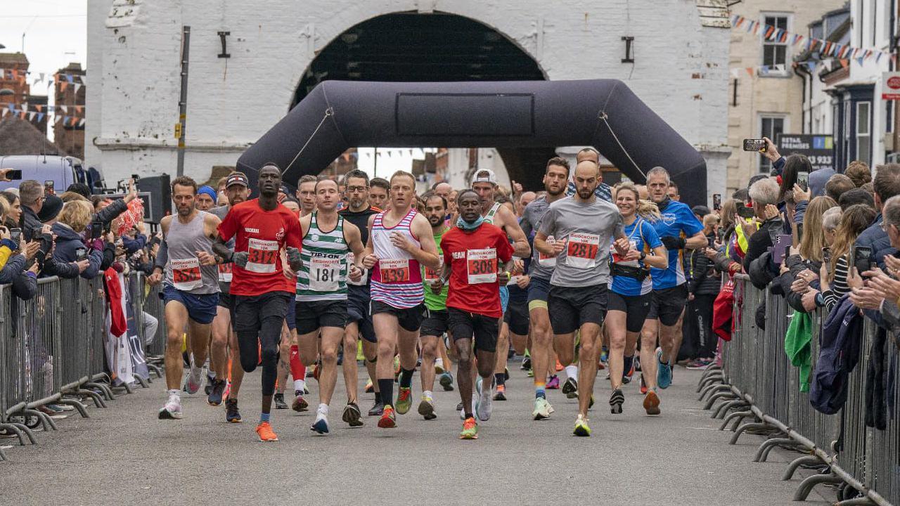 Lead runners start a race, wearing t-shirts, shorts and race numbers. An inflatable starting arch can be seen behind them and supporters behind metal barriers line the streets on both sides, holding up phone cameras.