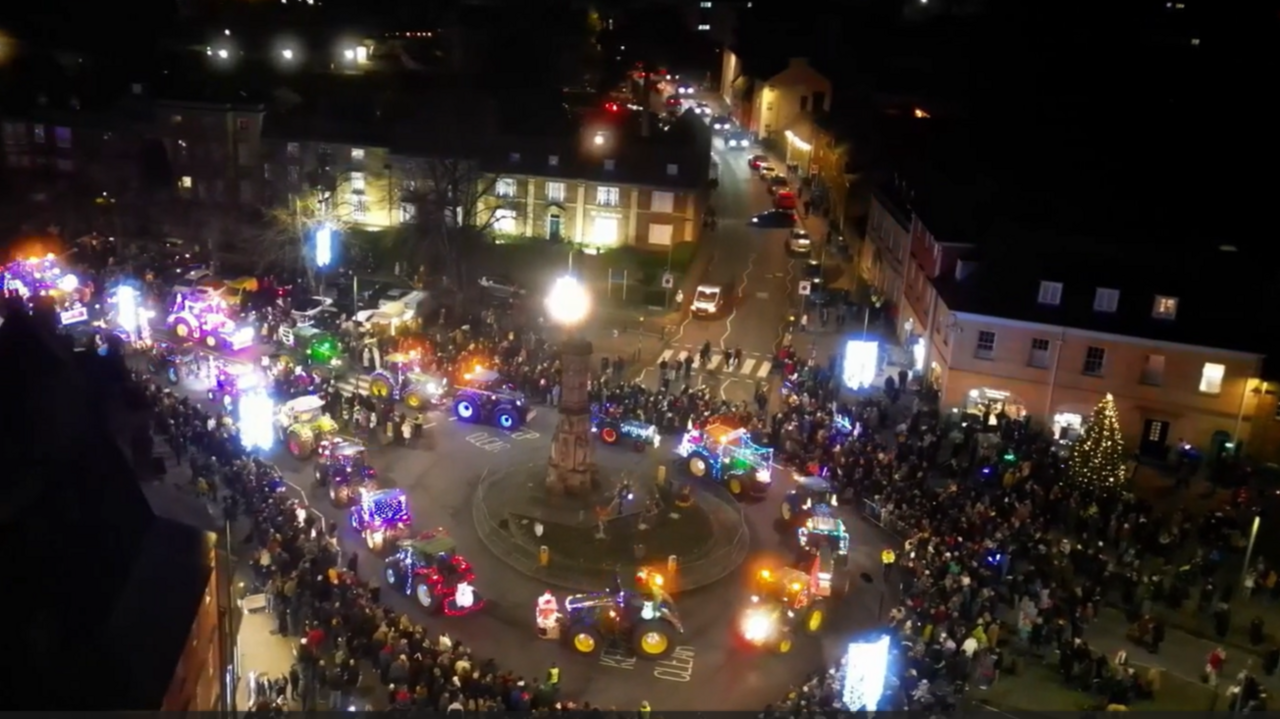 An aerial image of brightly lit tractors circling a square in front of crowds at night