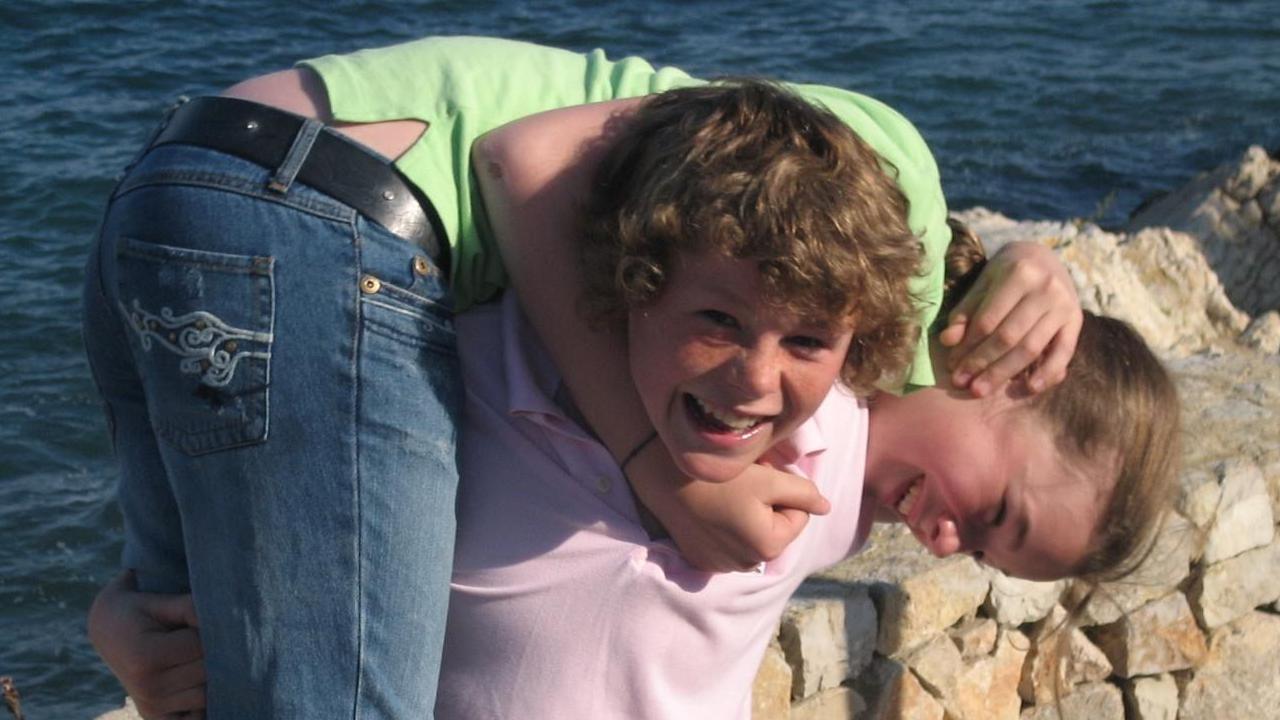 Charlotte being carried by Ryan as children. He is wearing a light pink polo top and she is wearing jeans and a green top. The sea is visible in the background over a light stone wall